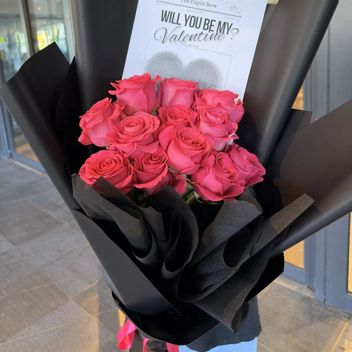 12 hot pink roses bouquet in black paper wrap with romantic gift card held outdoors, Sydney same-day luxury hot pink rose delivery by Vhouse Florist