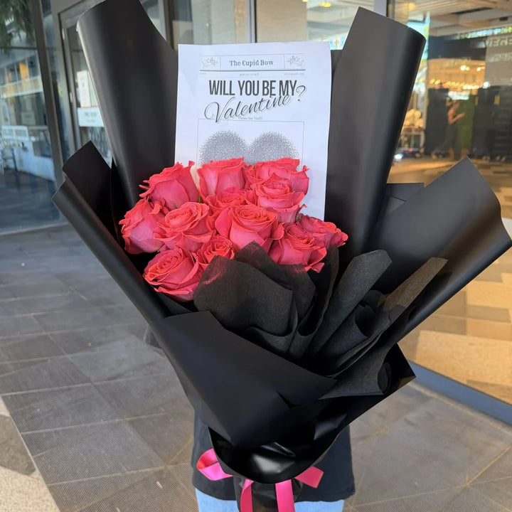 12 hot pink roses bouquet wrapped in black paper with pink ribbon and romantic gift card held outdoors, Sydney same-day luxury hot pink rose delivery by Vhouse Florist