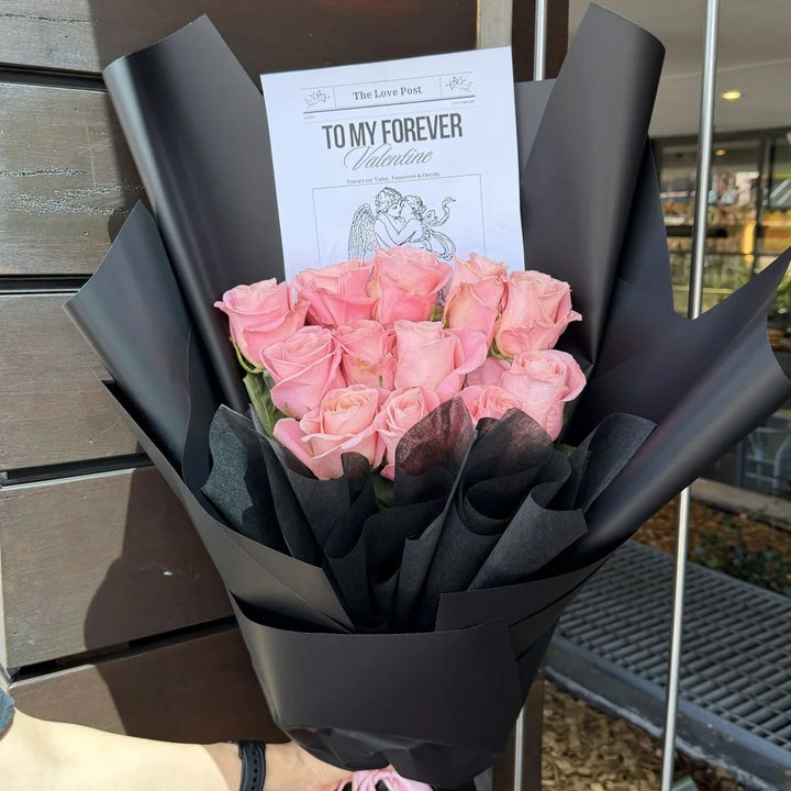 12 soft pink roses bouquet wrapped in black paper with romantic gift card held outdoors, Sydney same-day luxury pink rose delivery and romantic flower gifts by Vhouse Florist