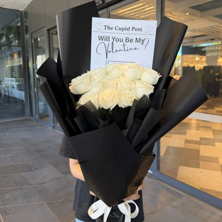 12 white roses bouquet wrapped in black paper with white ribbon and romantic gift card held outdoors, Sydney same-day luxury white rose delivery by Vhouse Florist