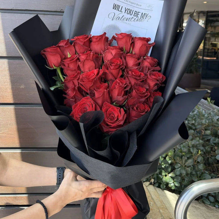 Person holding 24 luxury red roses bouquet wrapped in black paper with red ribbon and romantic gift card, Sydney same-day luxury red rose delivery by Vhouse Florist