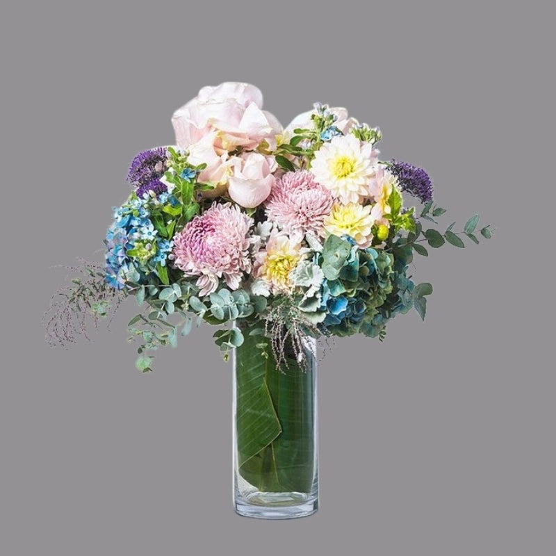 Luxury bouquet of hydrangea, soft pink roses, dusty miller, ast foliage, and texture in cylindrical glass vase.