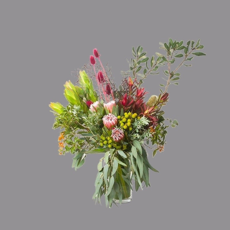 Native flowers like proteas, banksias, red thistle, wattle, and gum in cylindrical glass vase.