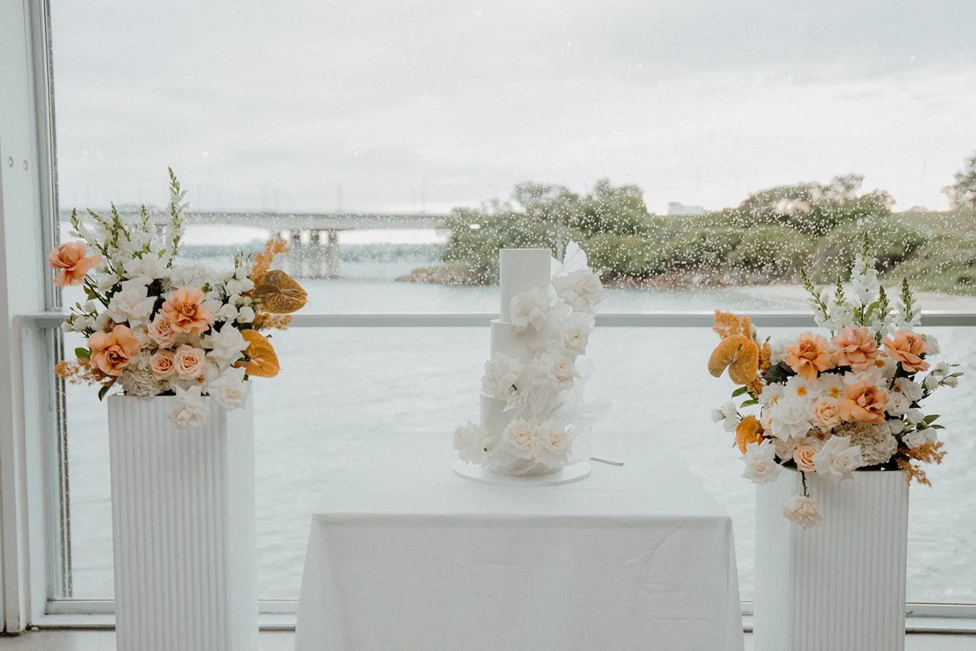 Waterfront wedding cake table flanked by apricot and ivory plinth flower arrangements, Sydney wedding flowers by Vhouse Florist