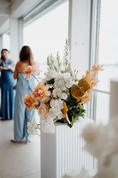Wedding reception entrance flowers on a white fluted plinth with peach roses and orange accents by a waterfront window